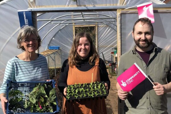 Three people smile while holding trays of seedlings and a Community Fund flag in front of a polytunnel