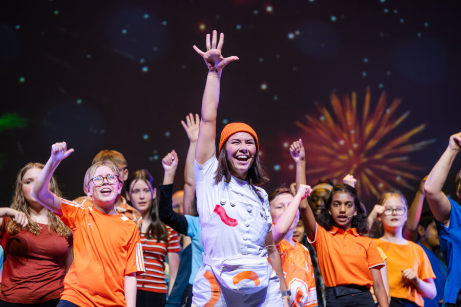 A young performer in an orange hat leads a group of children in a lively stage finale.
