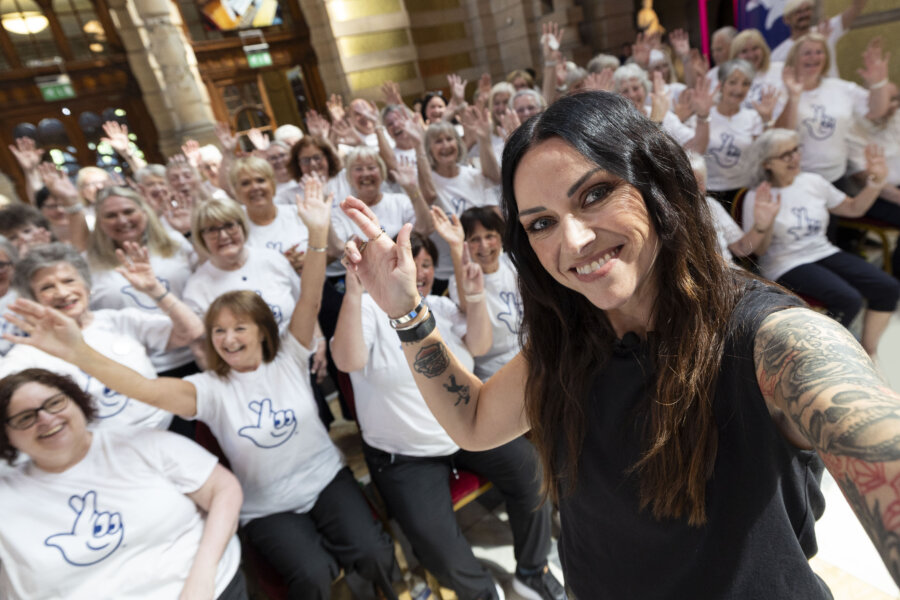 A woman with tattoos is taking a cross finger selfie. There is a group of people in the background who are smiling to the camera and wearing matching t-shirts.