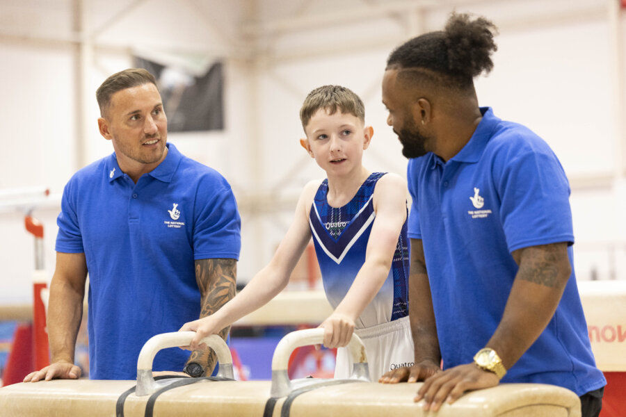 Two men in blue National Lottery-branded t-shirts talk to a young gymnast inside a gym.