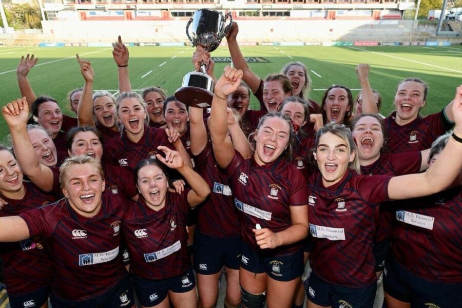 A rugby team in matching red jerseys carrying a trophy and cheering.