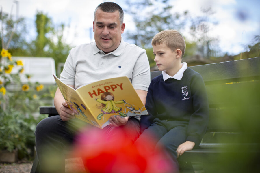 An older man and a young boy are sat outside on a bench with a book.