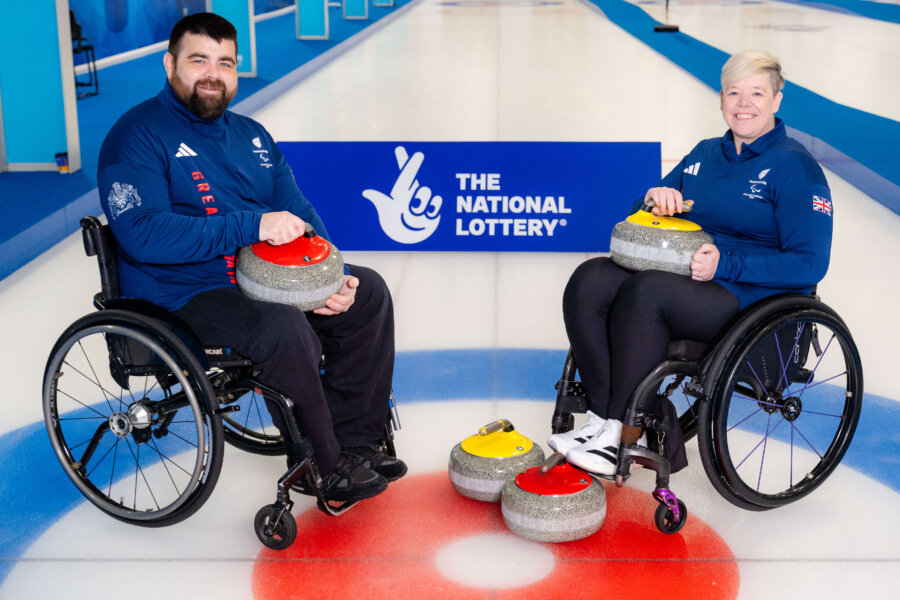 Two people in wheelchairs holding equipment used for curling and smiling at the camera.