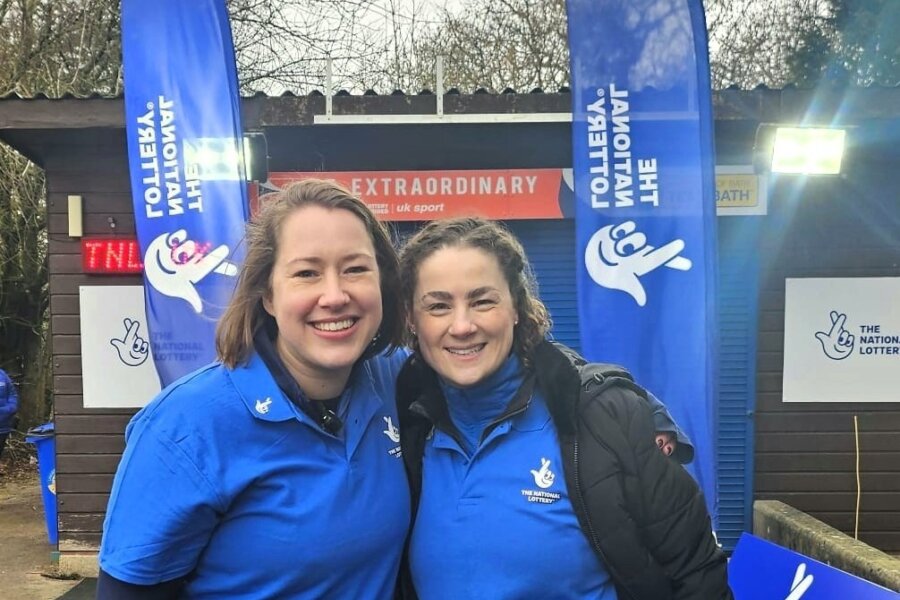 Two women wearing National Lottery branded tops, standing in front of National Lottery banners.