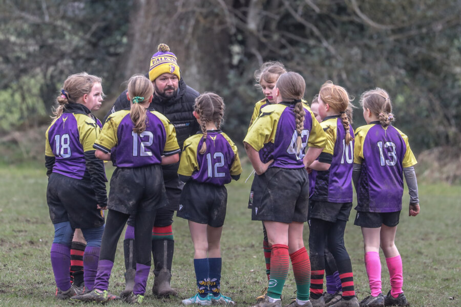 Young Rugby players in purple and yellow jerseys listen to a coach during a training session.