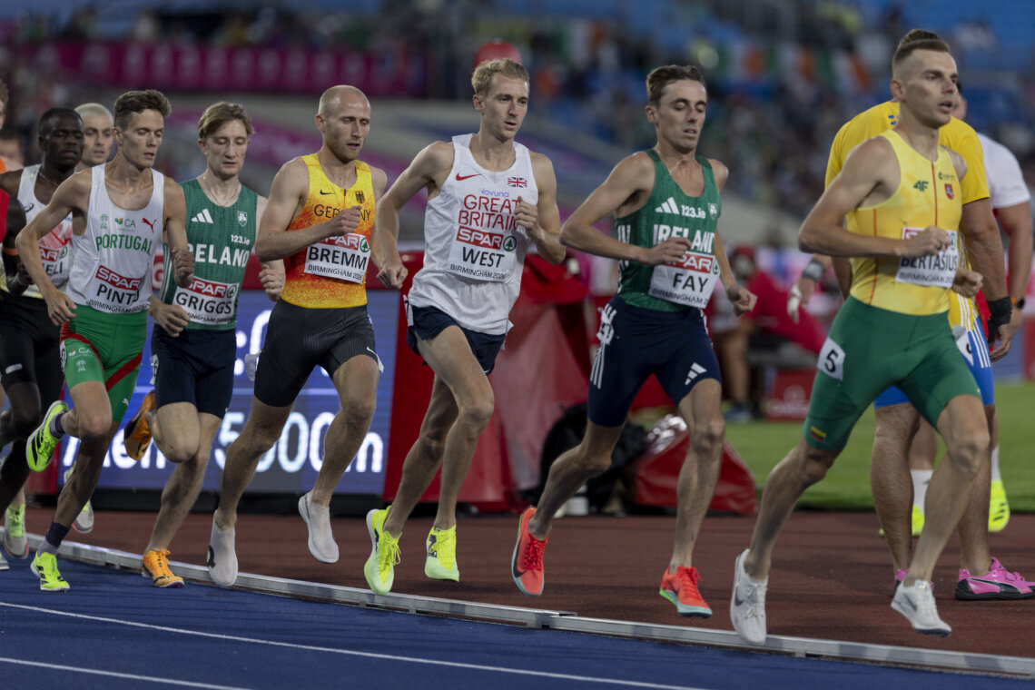 A group of male athletes in professional running kit race around a track.