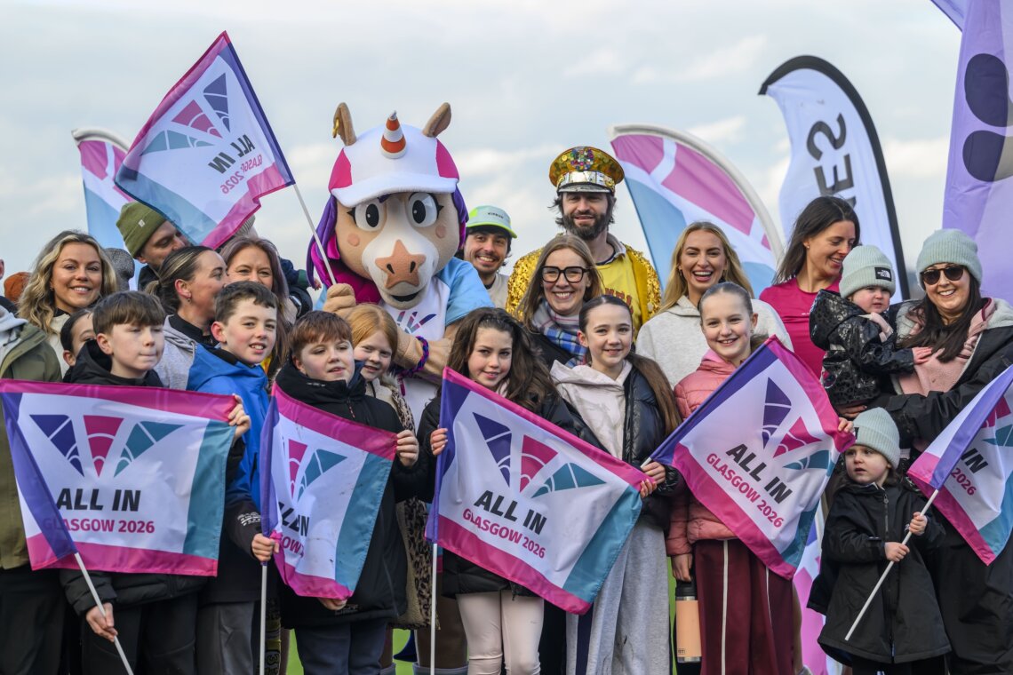 A group are gathered holding flags that say 'All in - Glasgow 2026'.