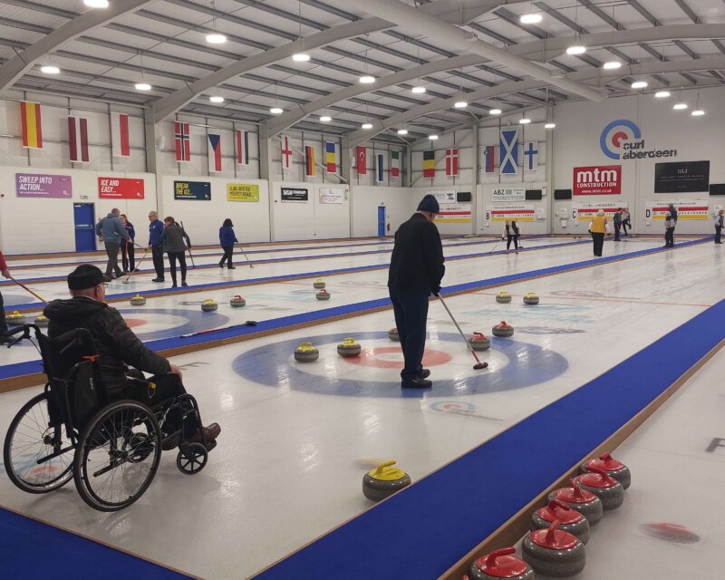 Wide view of the main ice hall at Curl Aberdeen, showing all six international-standard sheets in use. A wheelchair curler in the foreground reflects the club's fully inclusive offer, with members of all abilities competing together in the regular league. Image credit: Curl Aberdeen