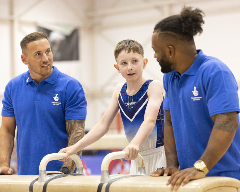 Two men in blue National Lottery-branded t-shirts talk to a young gymnast inside a gym.