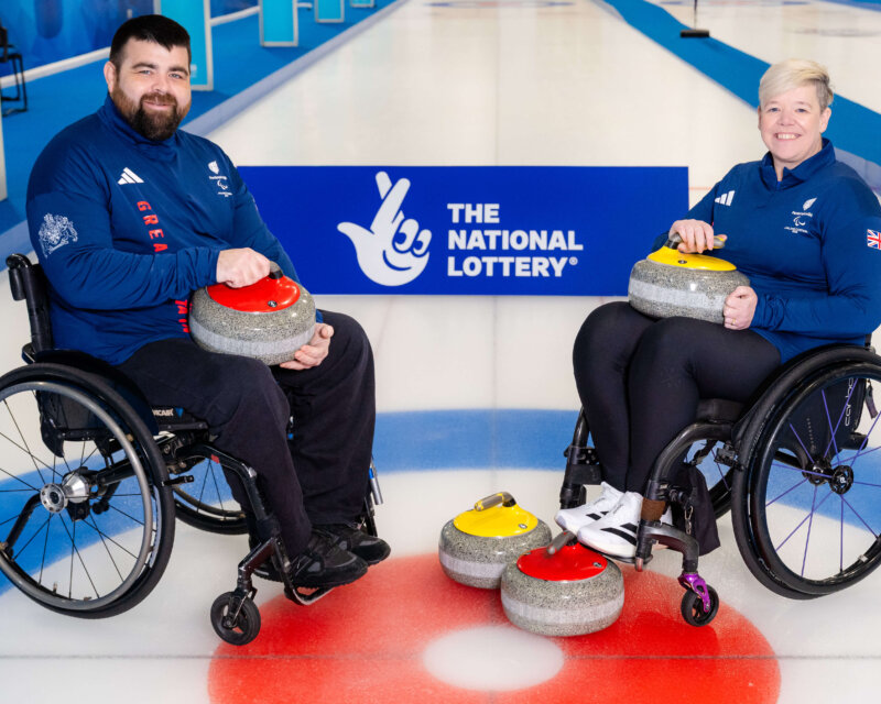 Two people in wheelchairs holding equipment used for curling and smiling at the camera.