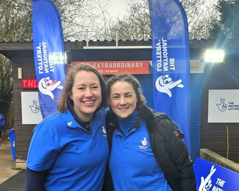 Two women wearing National Lottery branded tops, standing in front of National Lottery banners.