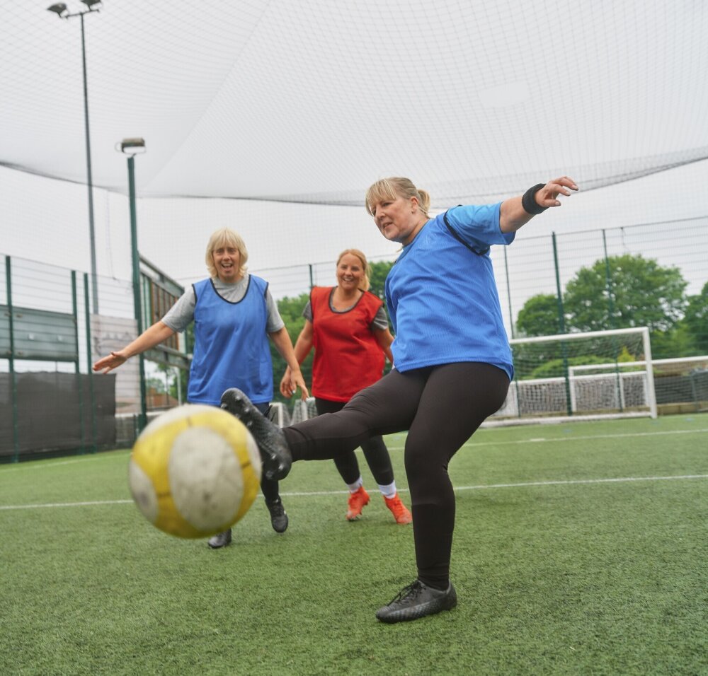Three women playing football on an outdoor pitch. One woman kicks the ball while two others watch and smile.