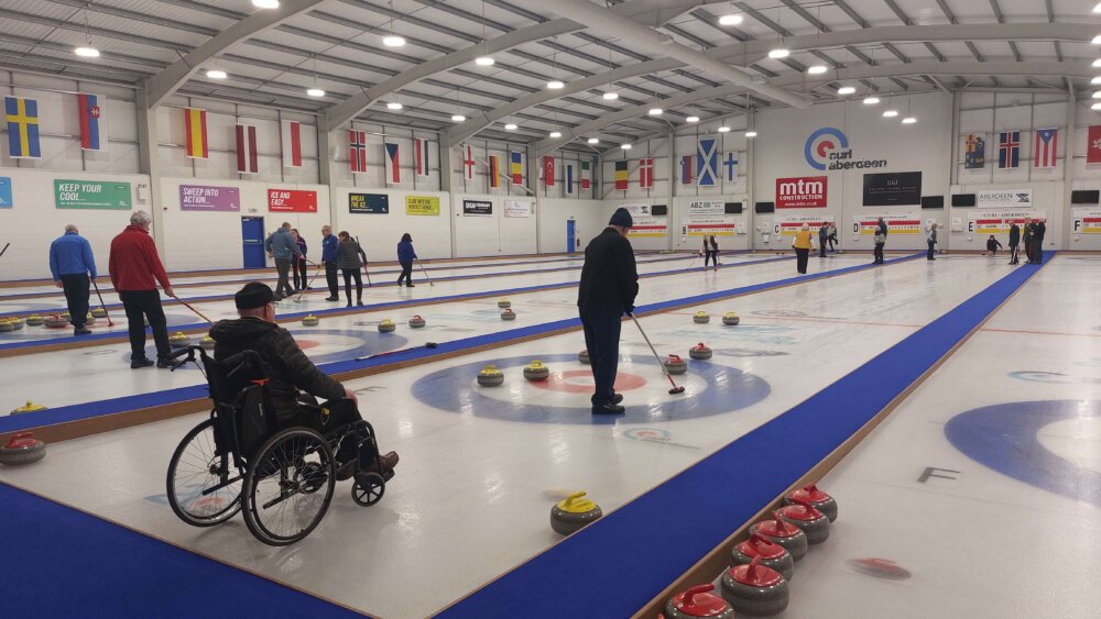 Wide view of the main ice hall at Curl Aberdeen, showing all six international-standard sheets in use. A wheelchair curler in the foreground reflects the club's fully inclusive offer, with members of all abilities competing together in the regular league.
