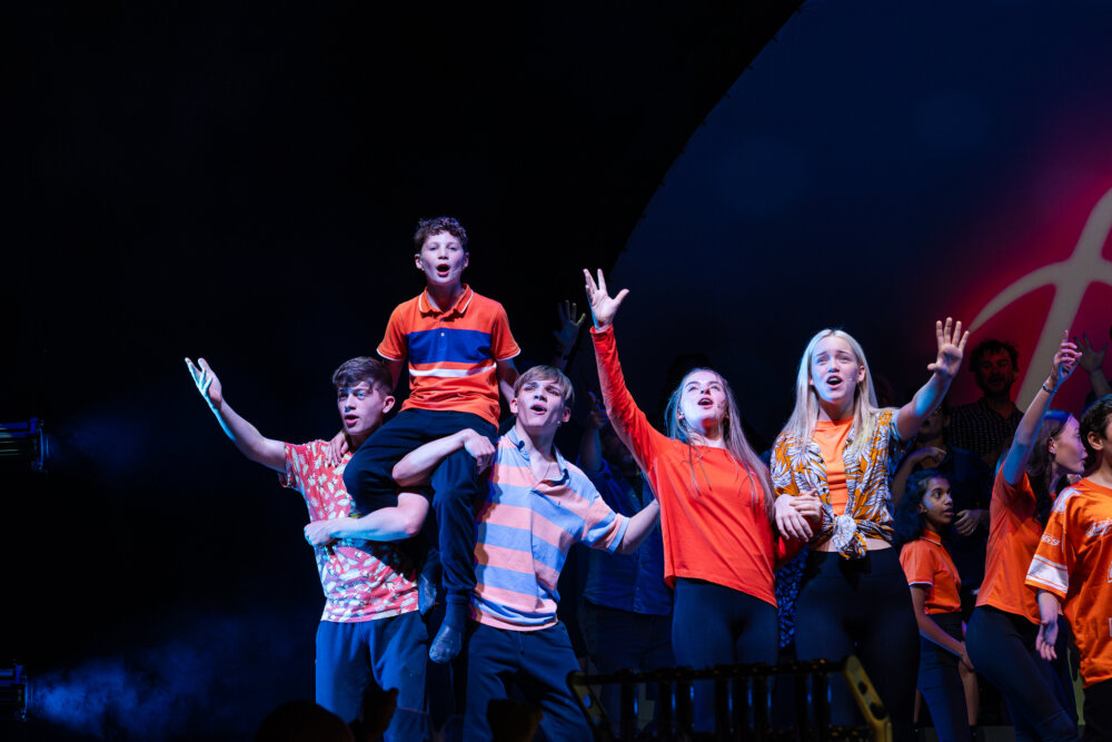A group of young performers sing on stage, with one child lifted on another’s shoulders.