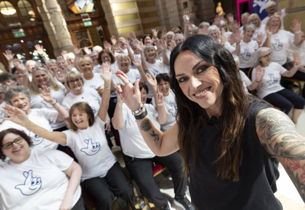 A woman with tattoos is taking a cross finger selfie. There is a group of people in the background who are smiling to the camera and wearing matching t-shirts.