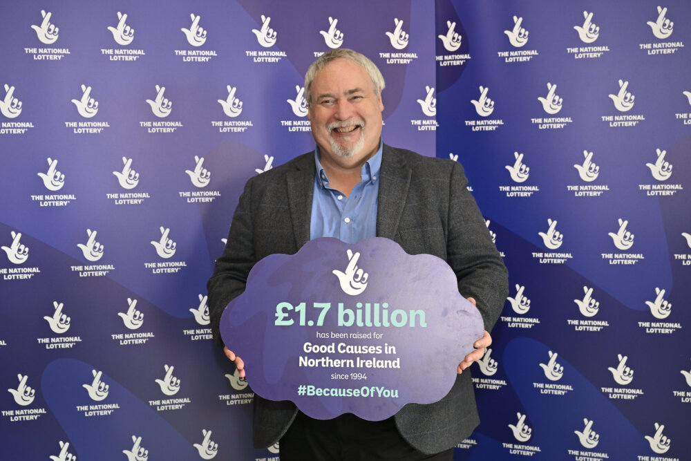 A person holds a sign with The National Lottery Logo in front of a branded banner.