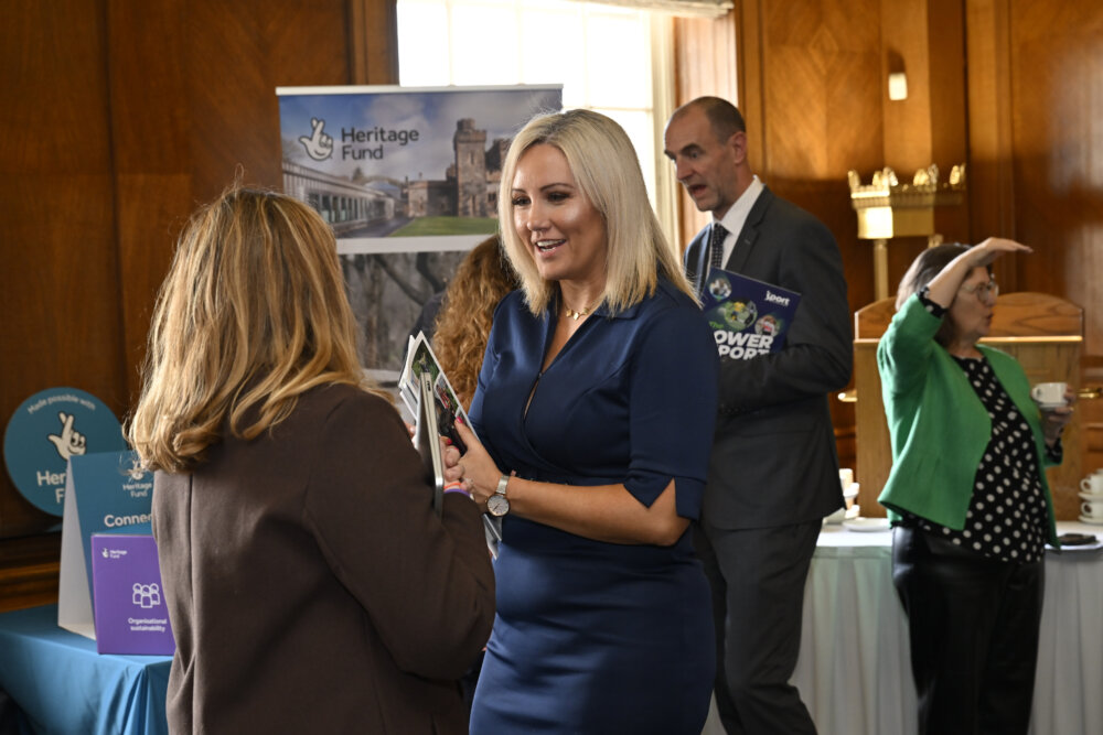 A smartly dressed woman in a hall holds documents while speaking to another woman.