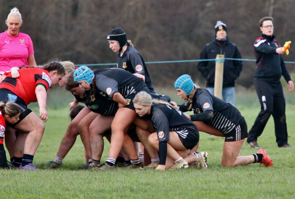 Girls rugby teams in black and red jerseys crouch into a scrum on a pitch.