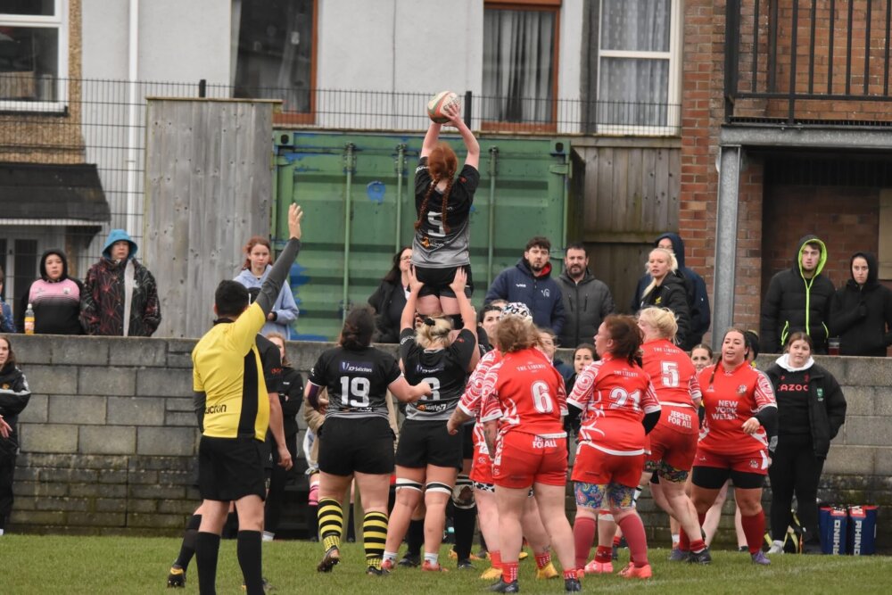 A women’s rugby match in progress, with a player in a black jersey lifted high by teammates to catch the ball.
