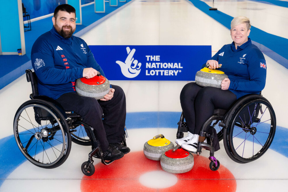 Two people in wheelchairs holding equipment used for curling and smiling at the camera.