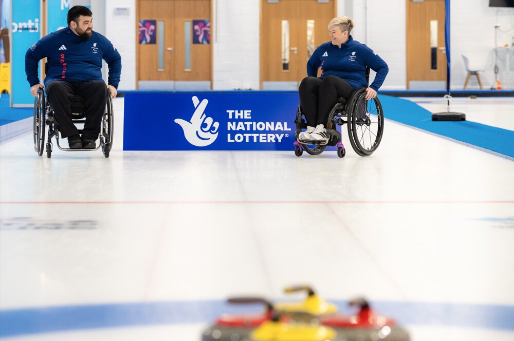 Two people in wheelchairs on the ice for curling.
