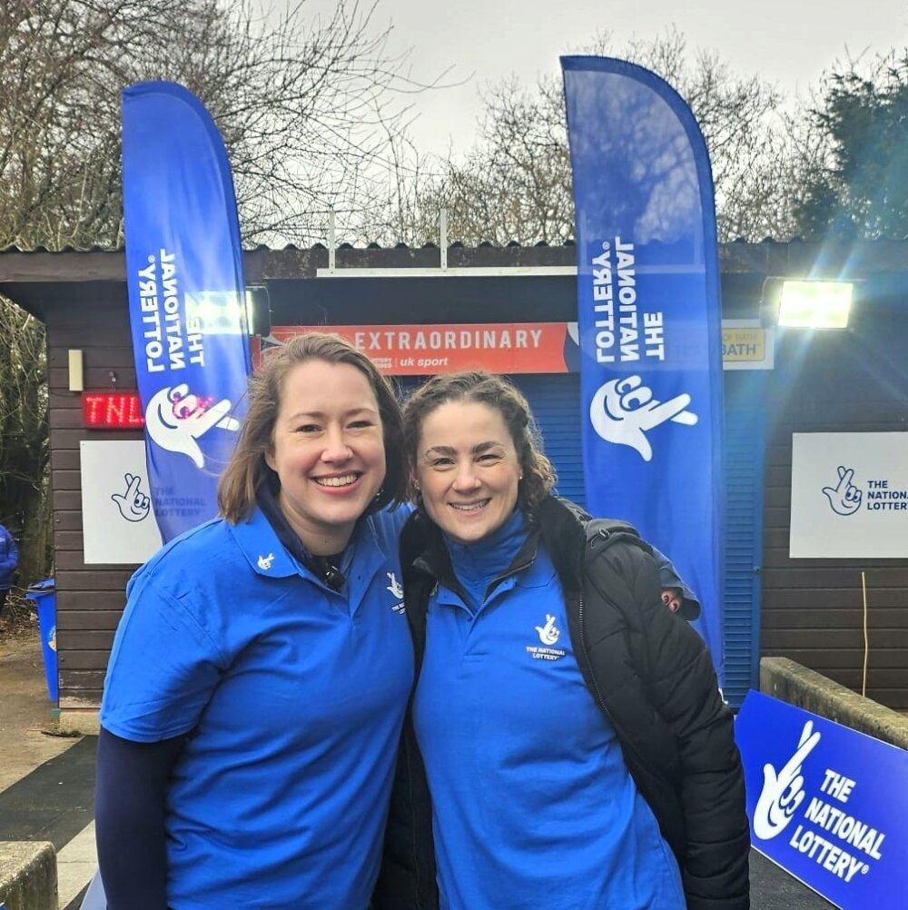 Two women wearing National Lottery branded tops, standing in front of National Lottery banners.