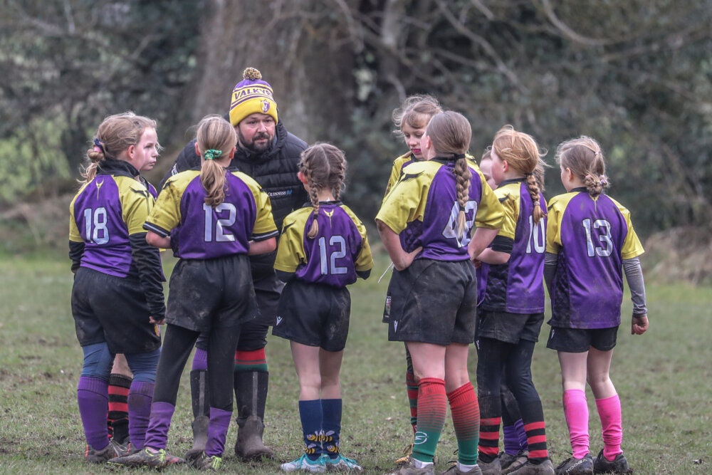 Young Rugby players in purple and yellow jerseys listen to a coach during a training session.