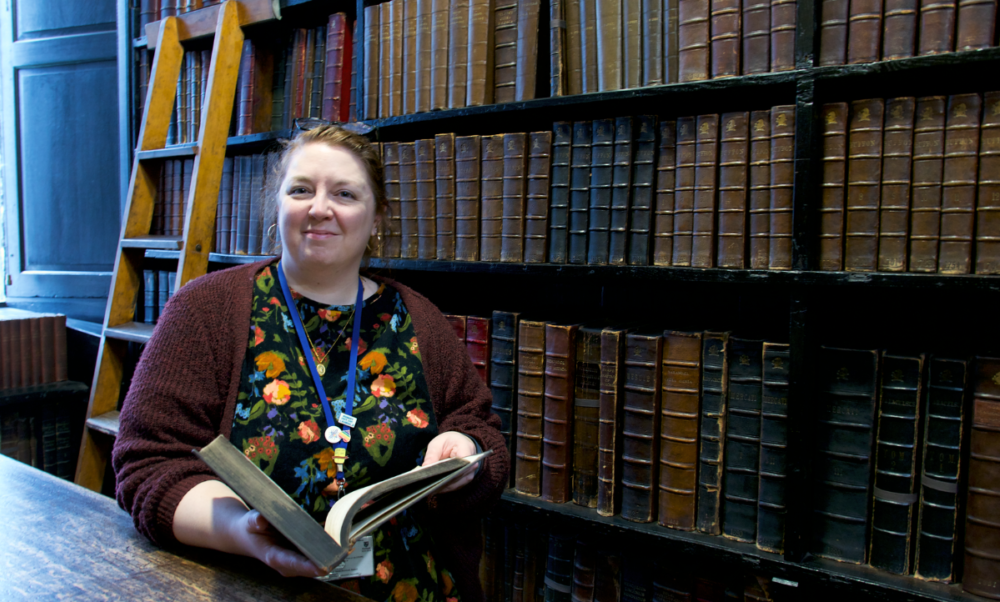 Photo showing Siân-Louise Mason, Visitor Services Coordinator, with a book in front of a book stack inside Chetham's Library.