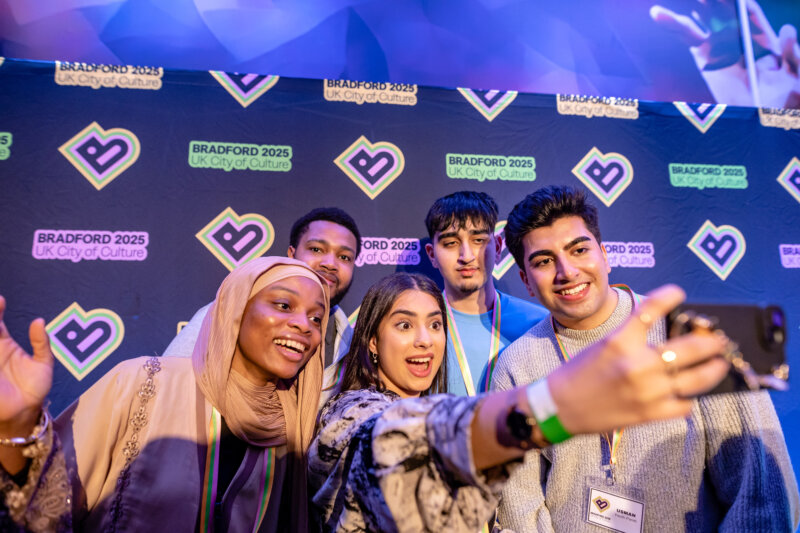A group of five smiling young people take a selfie together in front of a 'Bradford 2025 UK City of Culture' backdrop