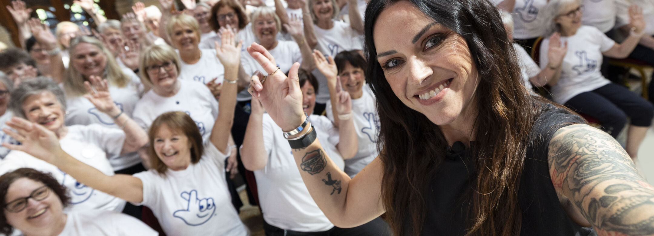 A woman with tattoos is taking a cross finger selfie. There is a group of people in the background who are smiling to the camera and wearing matching t-shirts.
