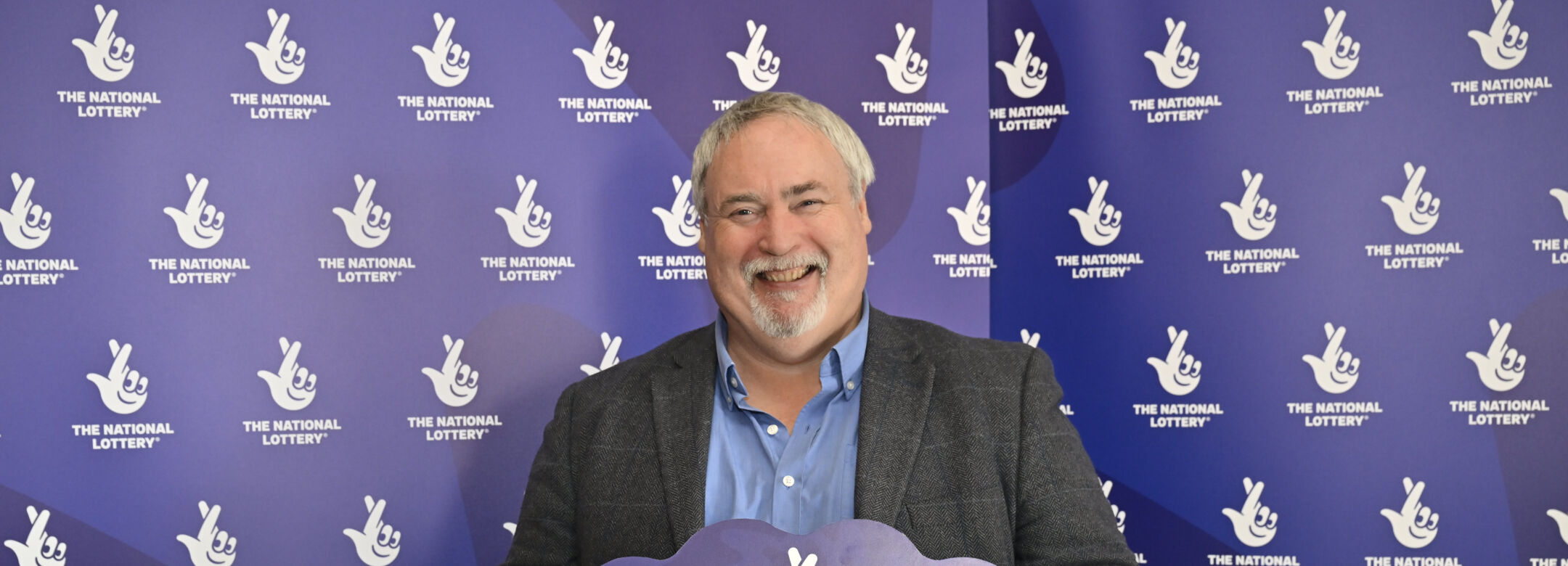 A person holds a sign with The National Lottery Logo in front of a branded banner.
