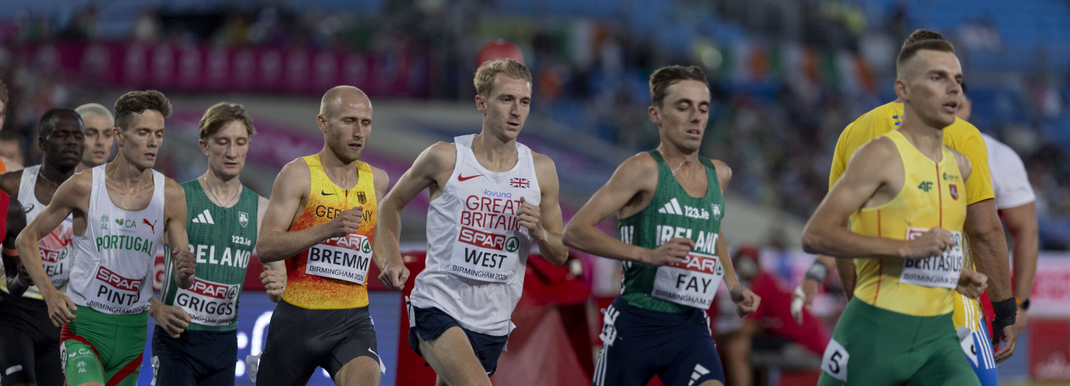 A group of male athletes in professional running kit race around a track.