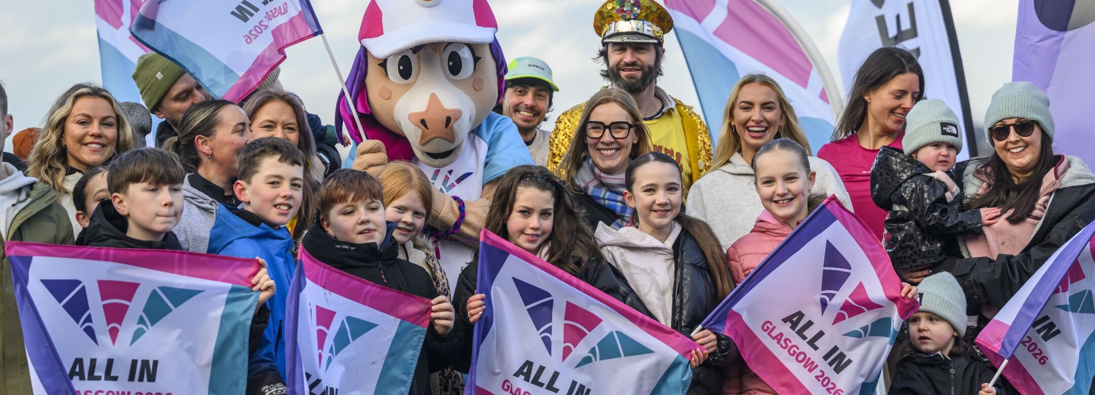 A group are gathered holding flags that say 'All in - Glasgow 2026'.