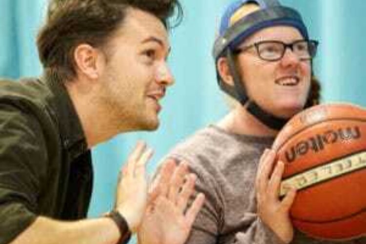 A young man is holding a basketball. He's sitting and about to throw it, and someone is next to him coaching him