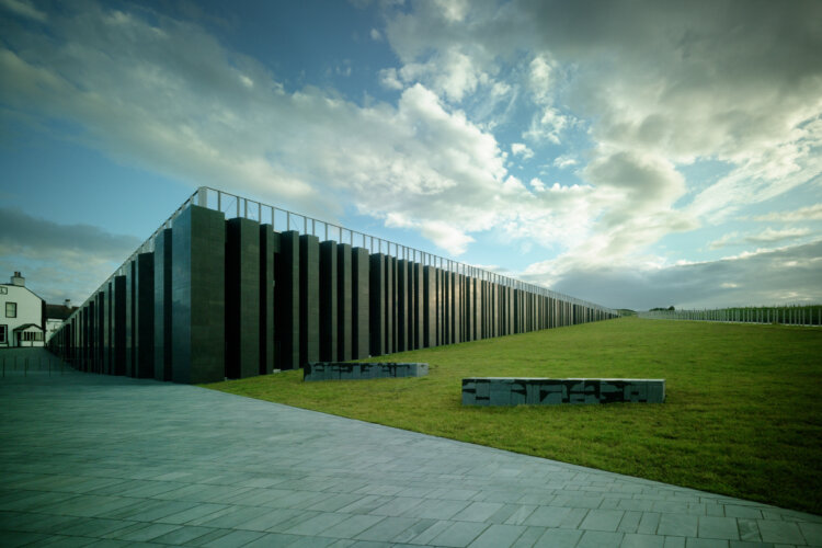 A large modern building with stone pillars. It is long and low under a vast cloudy sky