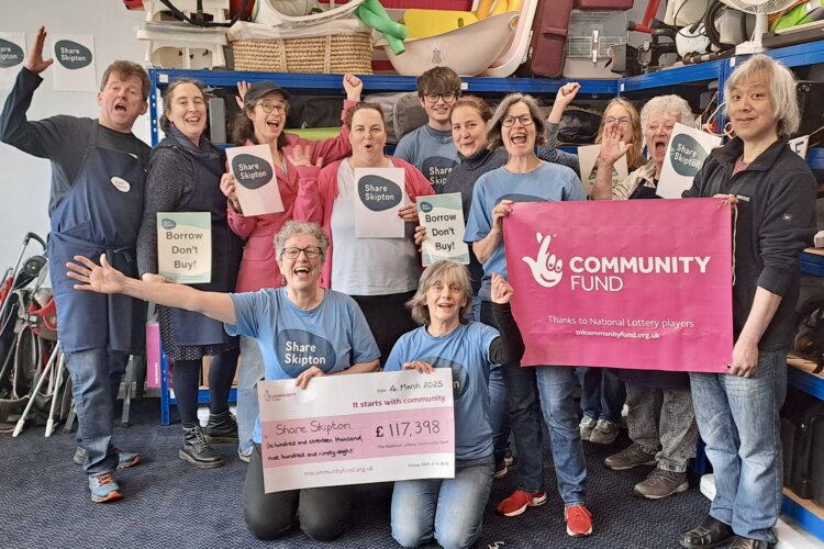 Photo shows an excited group of volunteers inside Share Skipton's library of things, holding a giant cheque, and celebrating their grant of £117,398 from the National Lottery Community Fund. Behind them are a variety of items available for lending.