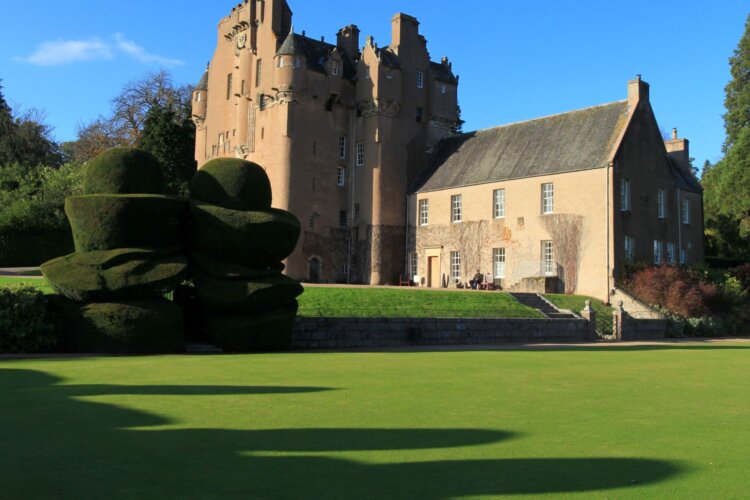 Large formal garden with neatly cut lawns, sculpted topiary, and surrounding trees under a bright blue sky.