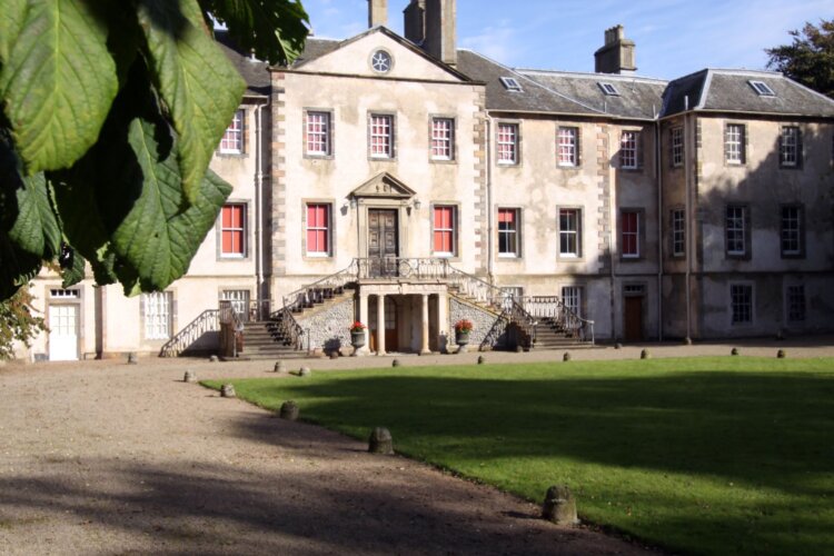 A large historic mansion with a symmetrical façade and central staircase, set behind an open lawn and framed by blossoming trees under a clear blue sky.
