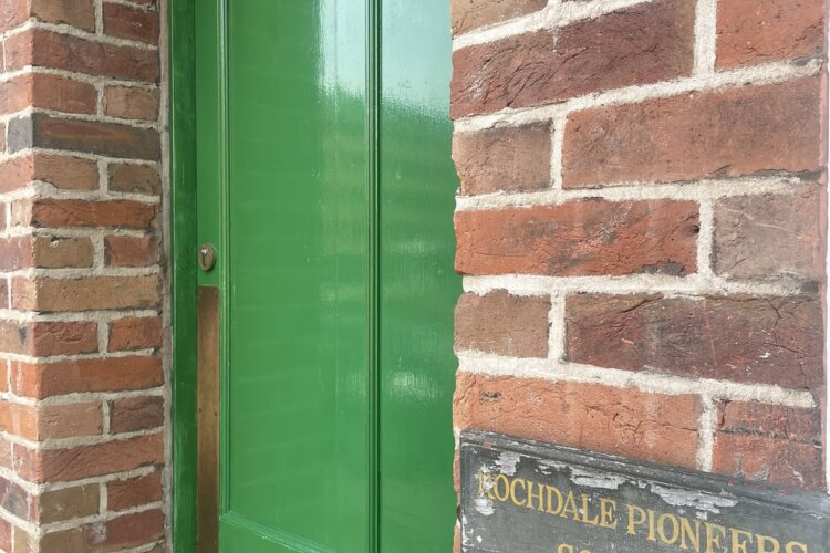 Colour image of a brick frontage with a green painted wooden door - which is the museum entrance. To the right of the door is a wooden hand painted sign which reads 'Rochdale Equitable Pioneers Society Est. 1844'