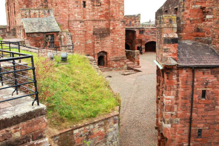 Ancient red-bricked castle courtyard with stone paths and arched entrances. Green grass grows on a raised section, and black metal railings line the walls.