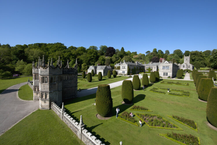 A grey brick gatehouse and country house beyond with visitors in the well manicured garden