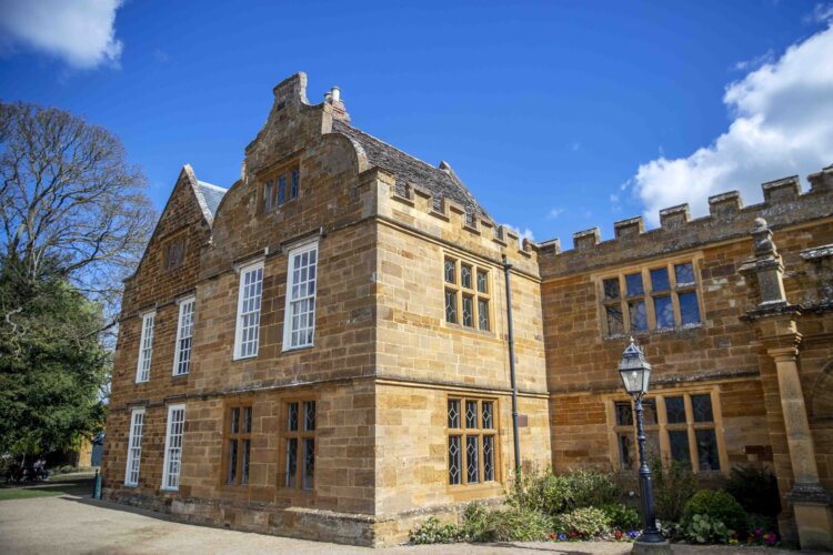 Image of part of the west range of Delapré Abbey during the daytime. It shows a portion of the building with large windows to the right, and a small portion of the parkland to the left.