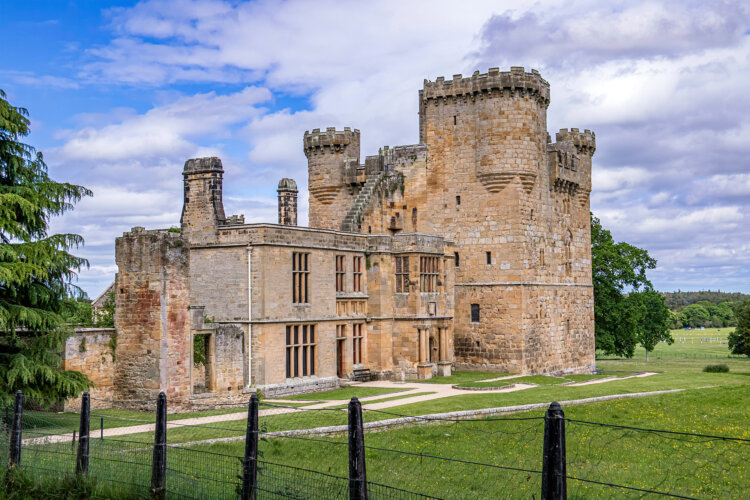 A historic stone castle with large rectangular windows and a round tower stands under a partly cloudy sky. It's surrounded by lush greenery and a fence.
