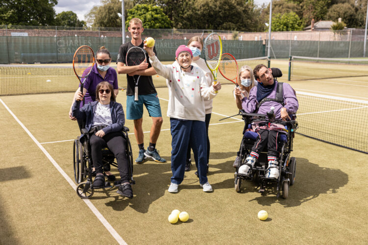 A group of 6 people are on a green tennis court holding tennis rackets in the air and smiling.
