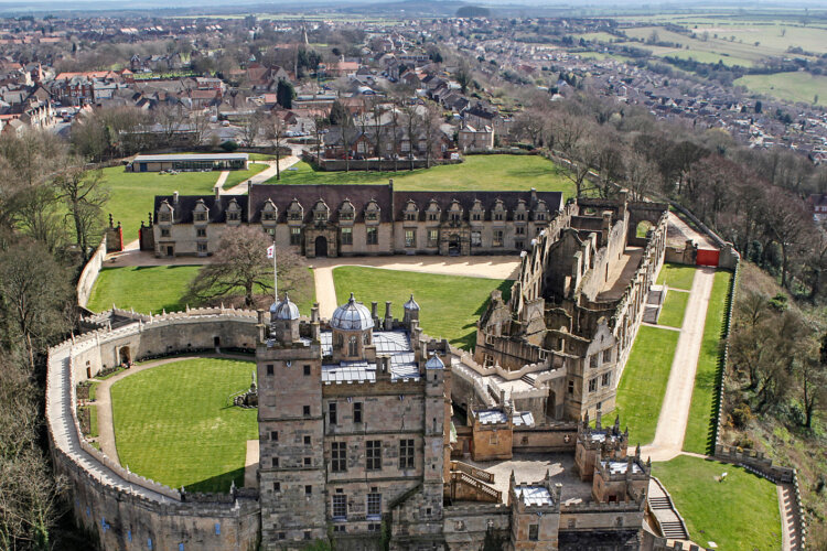 Aerial view of Bolsover Castle, a historic stone fortress with green lawns, surrounded by a town and countryside. The scene conveys a serene, timeless atmosphere.
