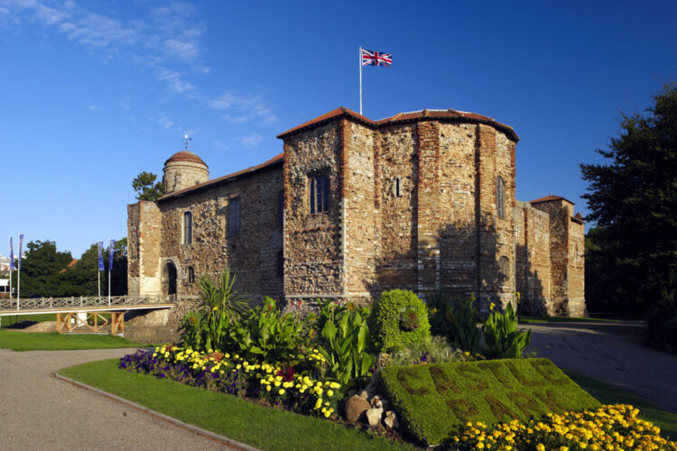 A photo of Colchester Castle, a large stone building surrounded by grass and footpaths. There is a flower bed in the foreground featuring yellow flowers. The sky is blue and clear.