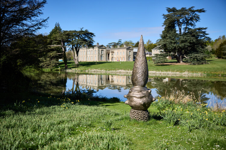 A bronze sculpture stands on grass beside a reflective lake, with Compton Verney house in the background, framed by mature trees under a clear blue sky.