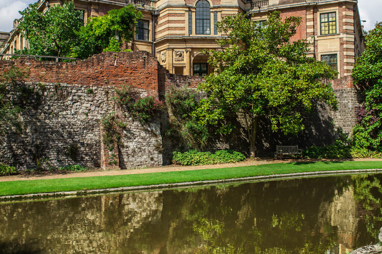A historic brick and stone building stands behind lush green trees and a serene pond. The scene conveys a peaceful, timeless atmosphere.
