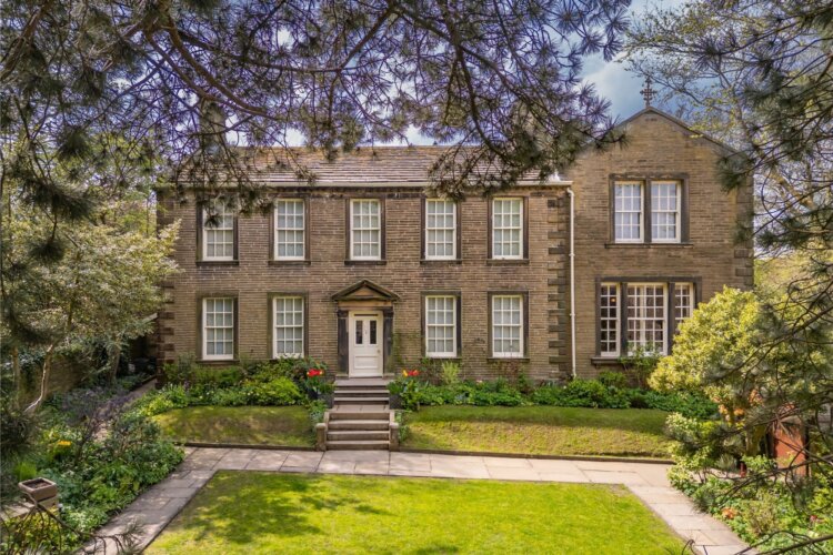 A brown stone building, the Brontë Parsonage Museum. It is set in a garden, with green bushes either side of a small stone staircase. The sky behind the building is a cloudy blue.