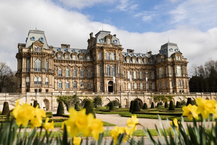 Image shows the exterior architecture of The Bowes Museum building.  The colour is a sandy stone effect.  Infront of the building are landscaped gardens and at the front of the image are yellow daffodils.